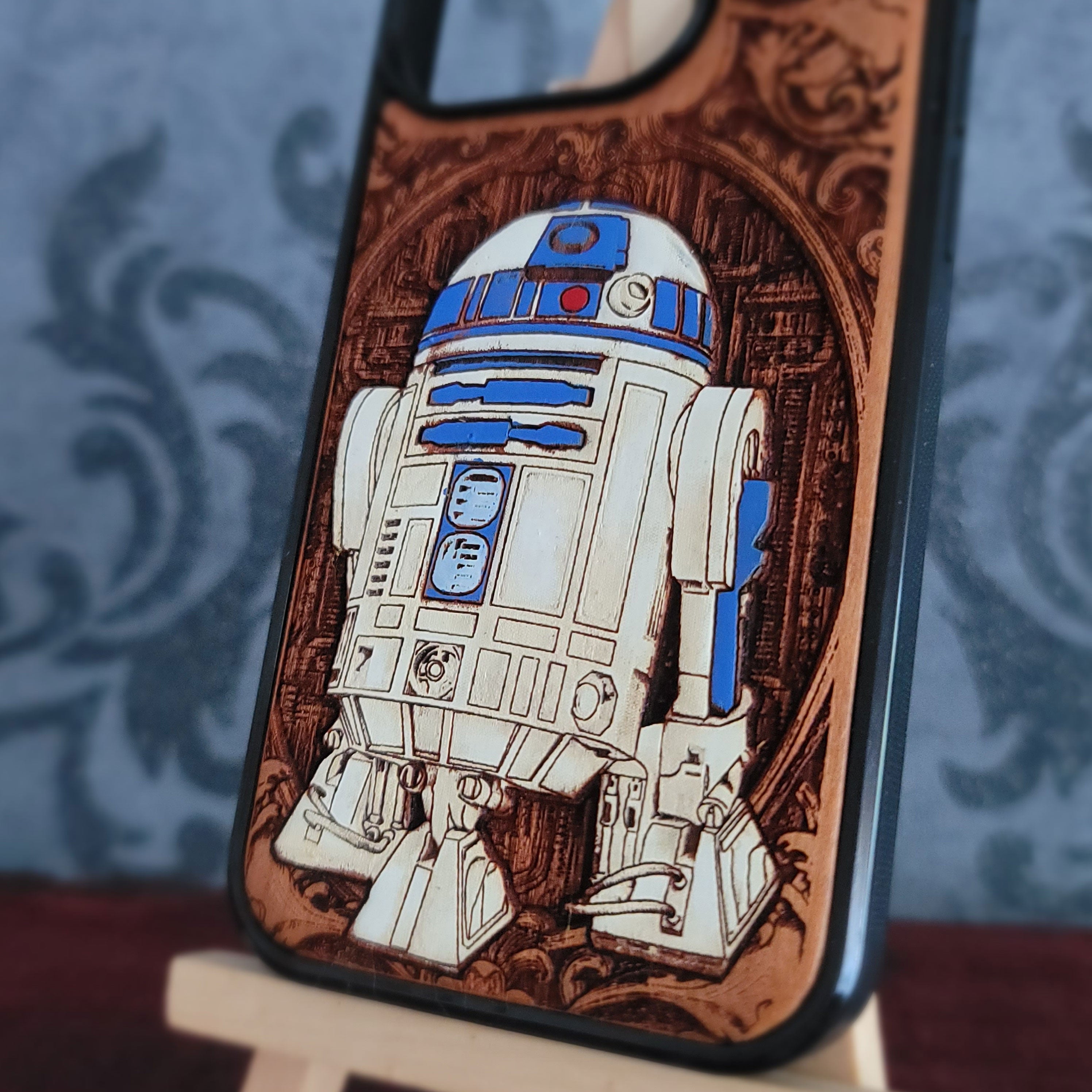 A close-up of a phone case featuring a detailed laser-burned design of the Star Wars droid R2-D2, set against a dark brown wood-grain pattern with a black protective bumper edge.