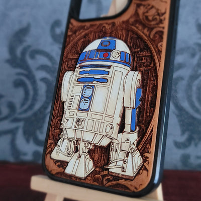A close-up of a phone case featuring a detailed laser-burned design of the Star Wars droid R2-D2, set against a dark brown wood-grain pattern with a black protective bumper edge.