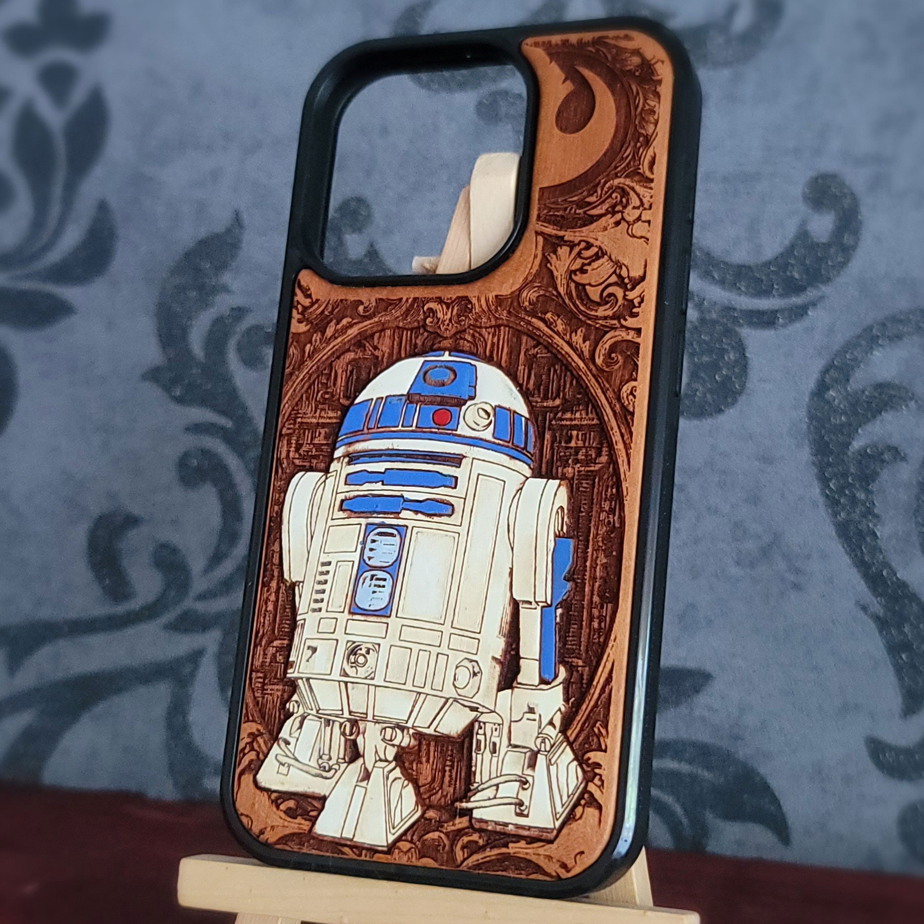 A custom wooden phone case featuring a detailed laser-engraved design of R2-D2 from Star Wars, displayed on a small wooden stand against a blue patterned background.