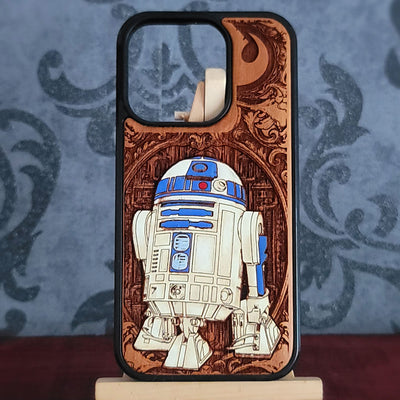 A wooden phone case featuring a laser-engraved Star Wars R2-D2 design, displayed on a small stand against a blue patterned background.