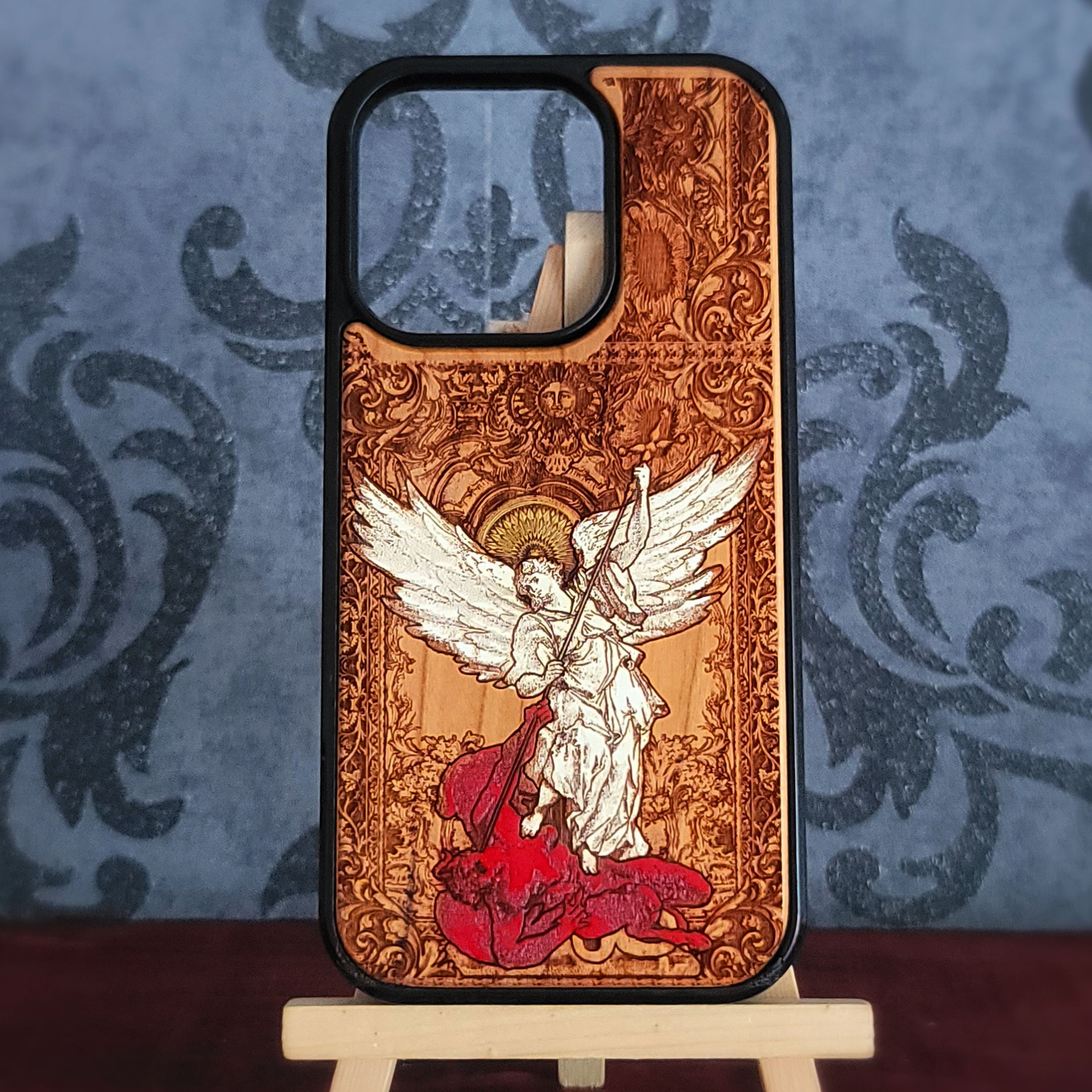 A wooden phone case with a black bumper edge, featuring a detailed laser-burned illustration of Saint Michael angel with white wings and a red cloak on a warm brown wood-grain background, displayed on a small wooden stand.