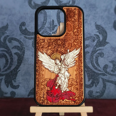 A wooden phone case with a black bumper edge, featuring a detailed laser-burned illustration of Saint Michael angel with white wings and a red cloak on a warm brown wood-grain background, displayed on a small wooden stand.