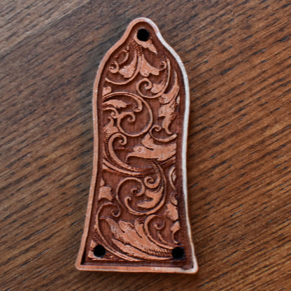 Laser‑engraved wood truss‑rod cover for an Epiphone‑style guitar. The wood has a medium brown finish with subtle grain texture. The central engraving features an elegant band of floral scrollwork. Two mounting holes are visible at the bottom edge.