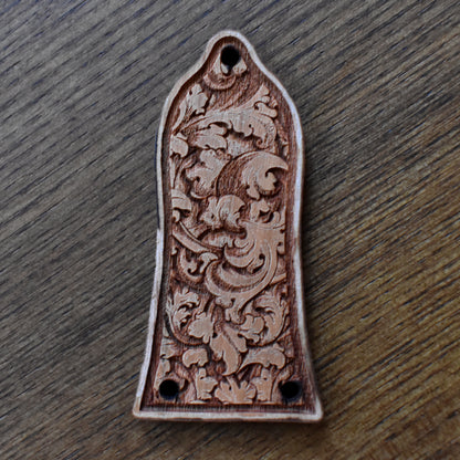 Laser‑engraved wood truss‑rod cover for an Epiphone‑style guitar. The wood has a light to medium brown finish with a faint figured grain. A band of delicate floral scrollwork spans the centre. The cover has two small mounting holes along the bottom edge