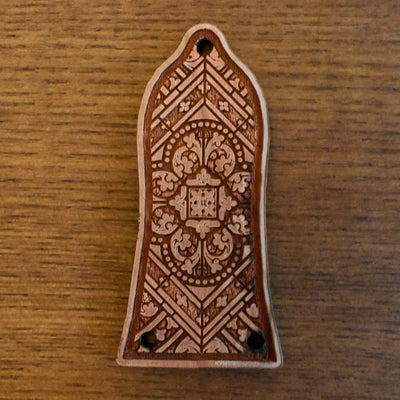 Laser‑engraved wood truss‑rod cover for an Epiphone‑style guitar. The wood has a light honey‑brown finish with a slightly figured grain. The engraving features a band of floral scrollwork extending horizontally across the centre, and two small mounting holes at the bottom edge.