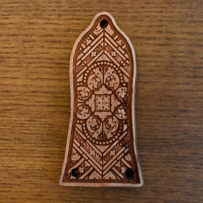 Laser‑engraved wood truss‑rod cover for an Epiphone‑style guitar. The wood has a light honey‑brown finish with a slightly figured grain. The engraving features a band of floral scrollwork extending horizontally across the centre, and two small mounting holes at the bottom edge.