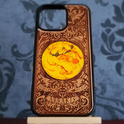 A custom wooden phone case with intricate laser-engraved baroque-style patterns, featuring a central circular yellow and orange marbled design and the name 'ARRAKIS' engraved at the bottom,