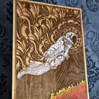 A wood burned artwork depicting an astronaut floating in space above a fiery red planet. The astronaut is wearing a white spacesuit and helmet. The background is a dark brown wood with intricate details.