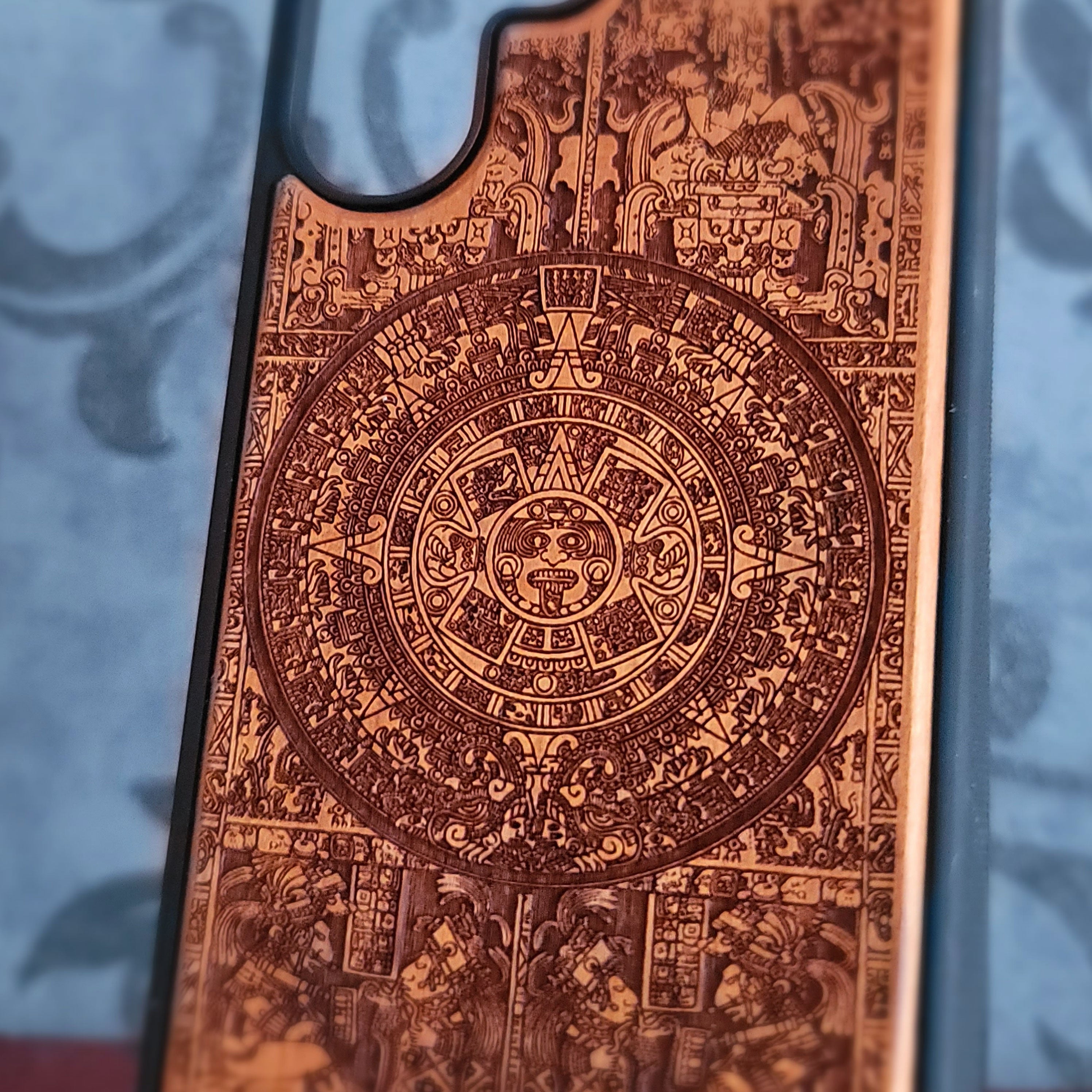 A close-up of a phone case featuring an intricate, detailed Aztec calendar design in warm copper-brown tones against a blurred blue-gray background.