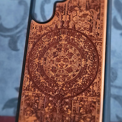 A close-up of a phone case featuring an intricate, detailed Aztec calendar design in warm copper-brown tones against a blurred blue-gray background.