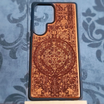 A phone case with a detailed, laser-engraved Aztec calendar design, featuring a warm copper-brown pattern on a black protective bumper, displayed on a blue patterned fabric background.