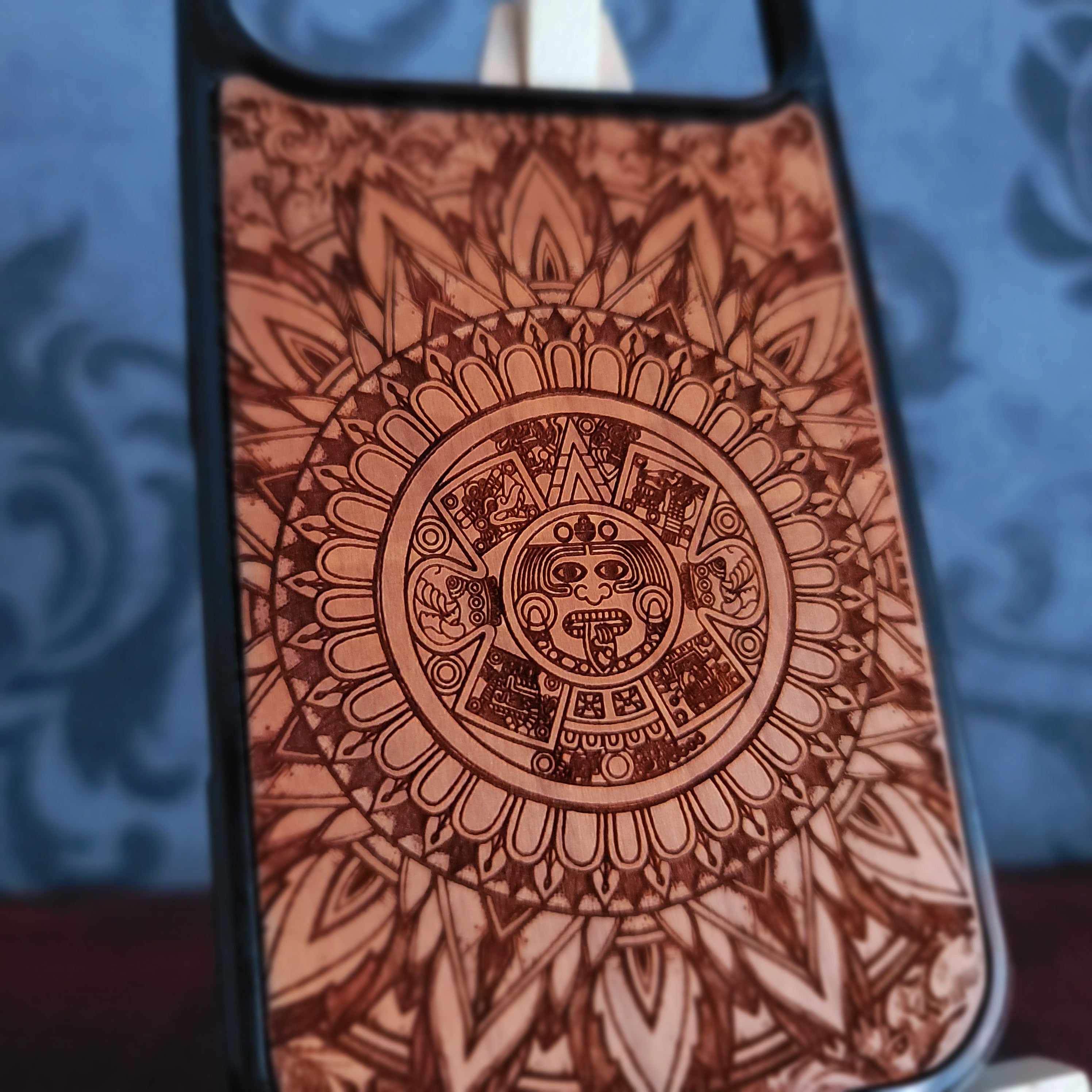 A close-up of a phone case featuring an intricate brown etched Aztec calendar design on a tan background, with a black bumper edge.