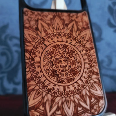 A close-up of a phone case featuring an intricate brown etched Aztec calendar design on a tan background, with a black bumper edge.