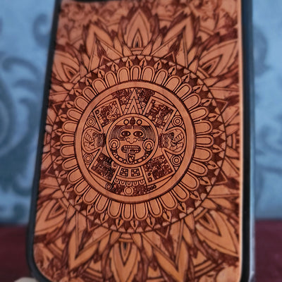 A close-up of a phone case featuring a detailed, circular Aztec calendar-style sun design in dark brown engravings over a warm orange-brown wood-like finish.