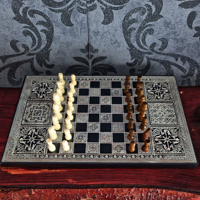 An ornate chess set displayed on a dark wooden table, featuring a black and silver patterned board with cream-colored light pawns on the left and brown dark pawns on the right, set against a gray damask patterned backdrop.
