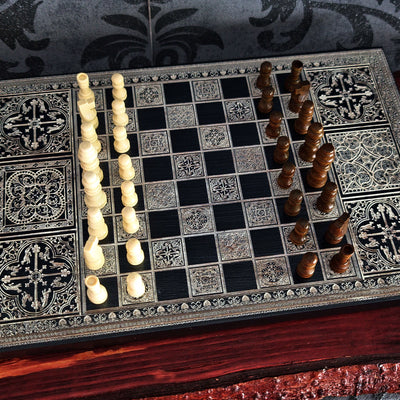 An ornate chess set displayed on a decorative chessboard. The board features black and silver-toned square tiles with intricate filigree patterns, accompanied by ivory and dark brown carved chess pieces arranged on opposite sides.