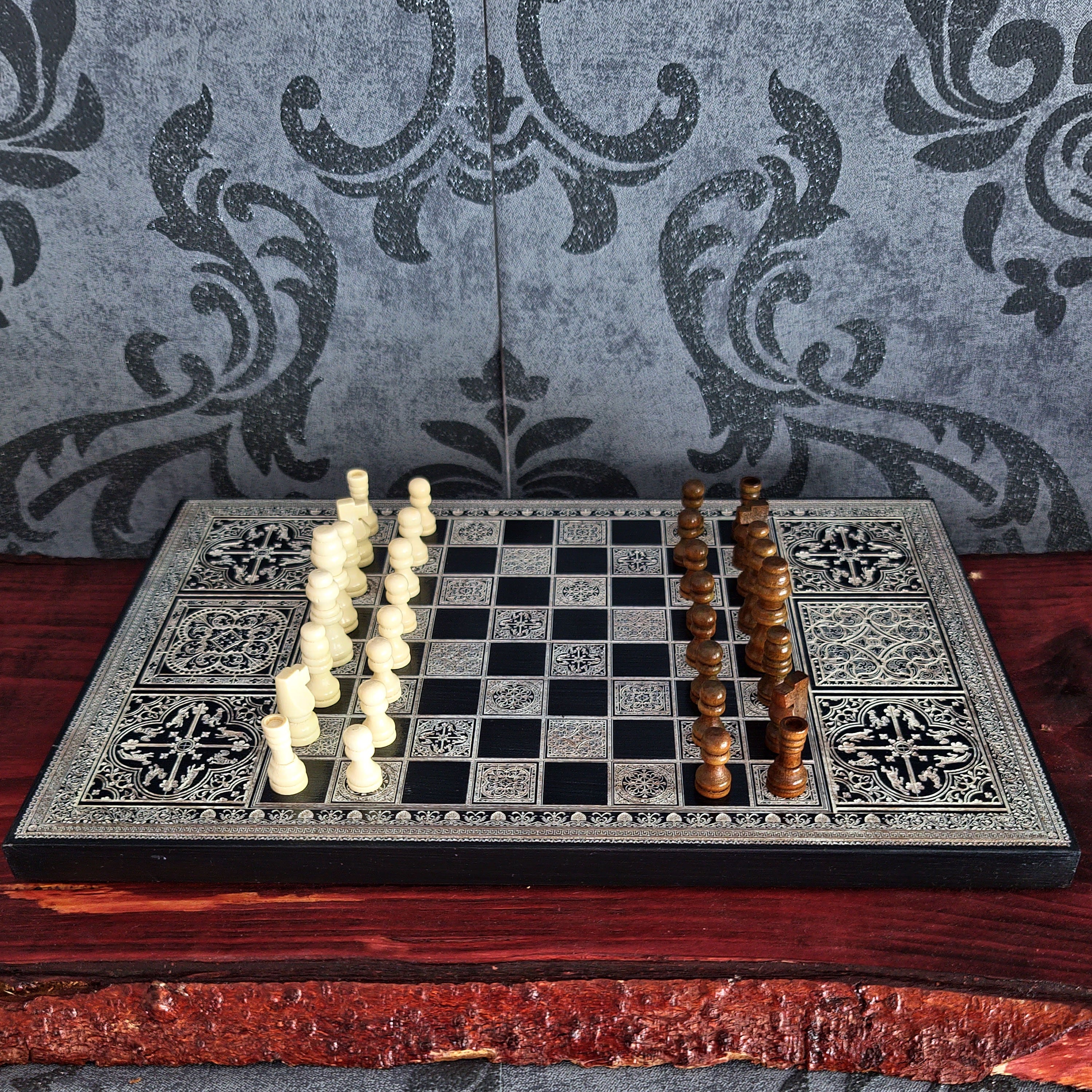 An ornate chess set with a black and silver patterned board, arranged on a dark wooden surface against a gray damask-patterned backdrop. The set includes ivory and brown wooden chess pieces.