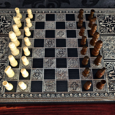An ornate chess set with a black and silver patterned board, featuring cream-colored pawns on one side and dark brown wooden pieces on the other, photographed from a top-down angle.