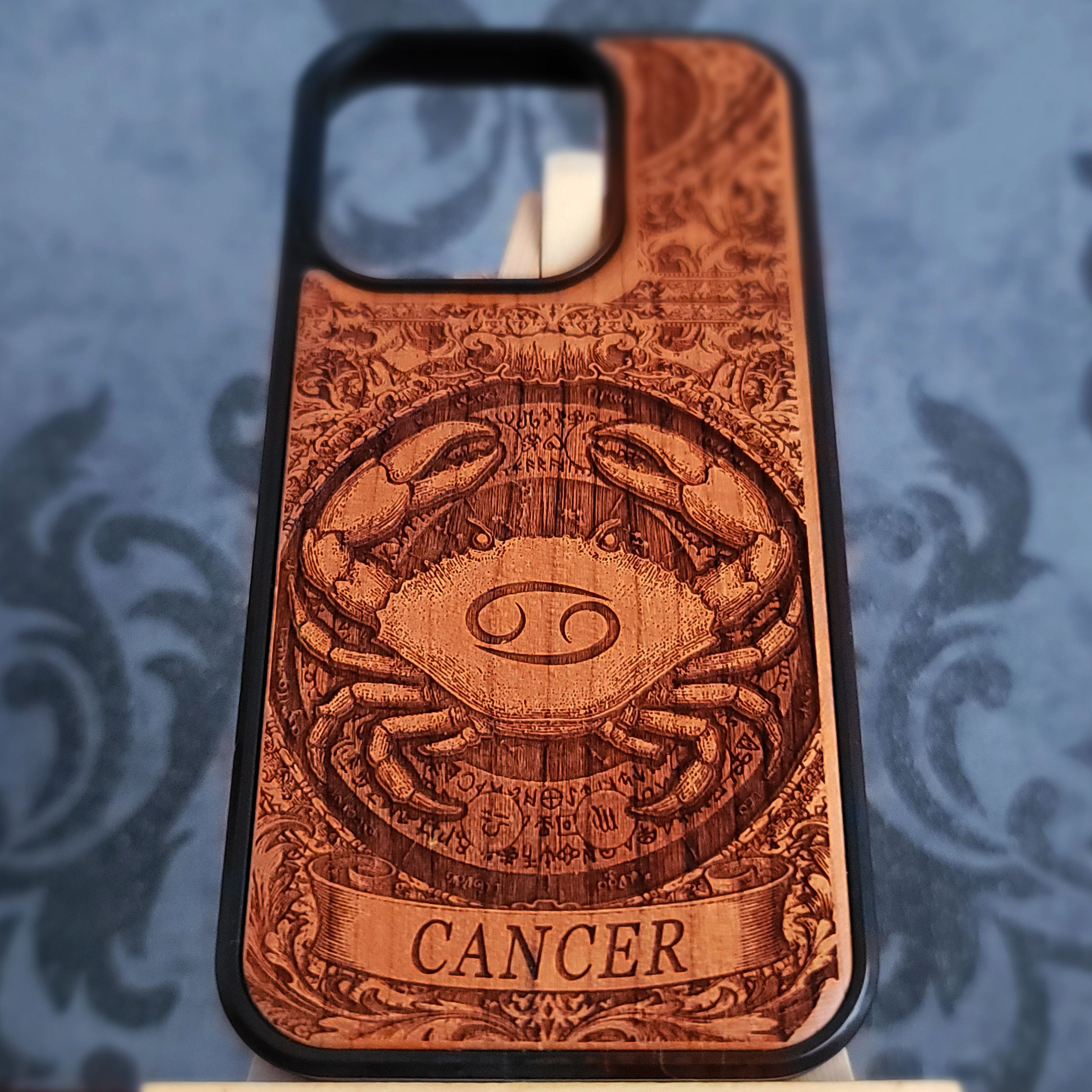 A wooden phone case featuring a detailed laser-engraved crab design representing the Cancer zodiac sign, with the word 'CANCER' on a banner at the bottom, set against a blue patterned background.