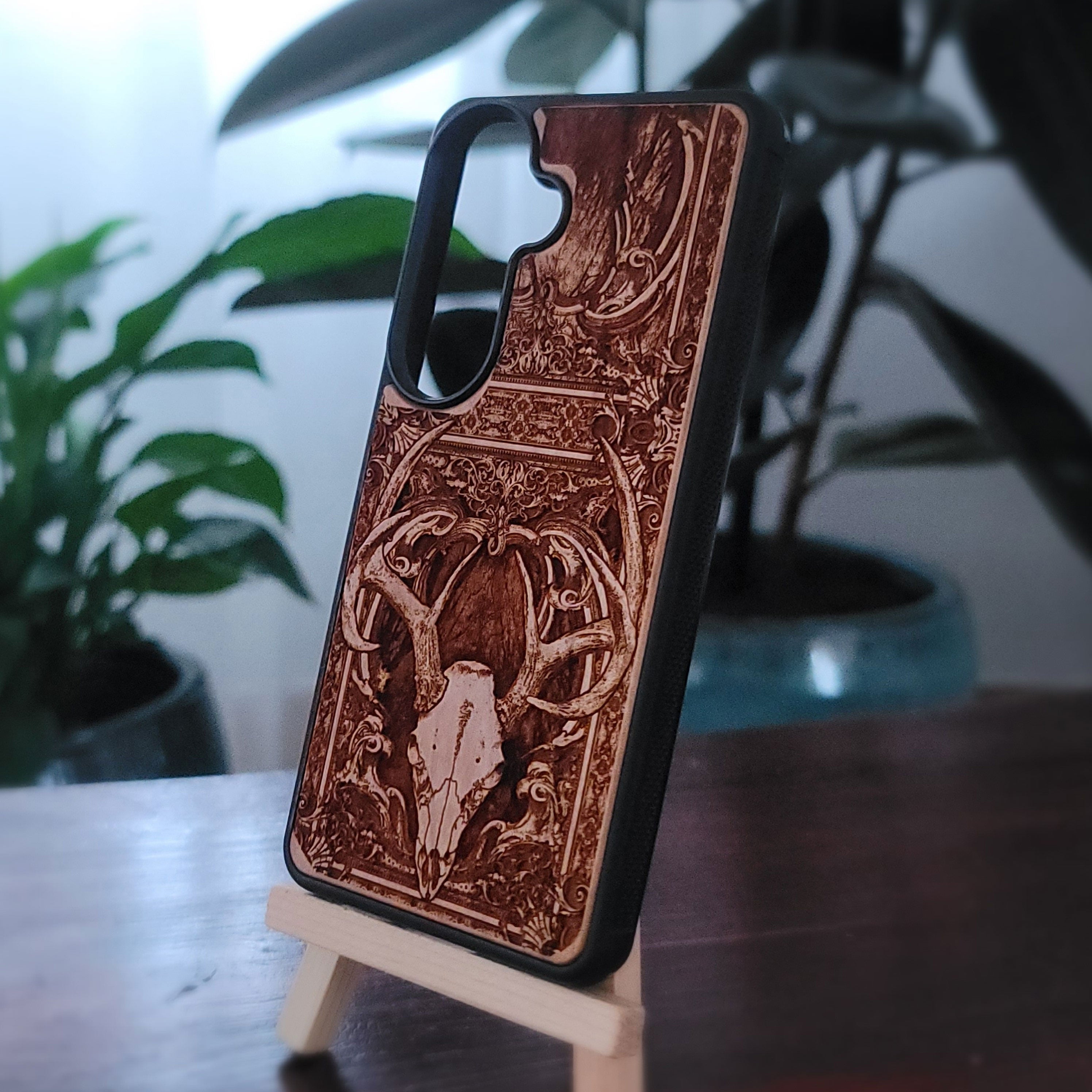 A wooden phone case with an engraved deer skull and antler design, displayed on a small wooden stand in an indoor setting with blurred plants in the background.