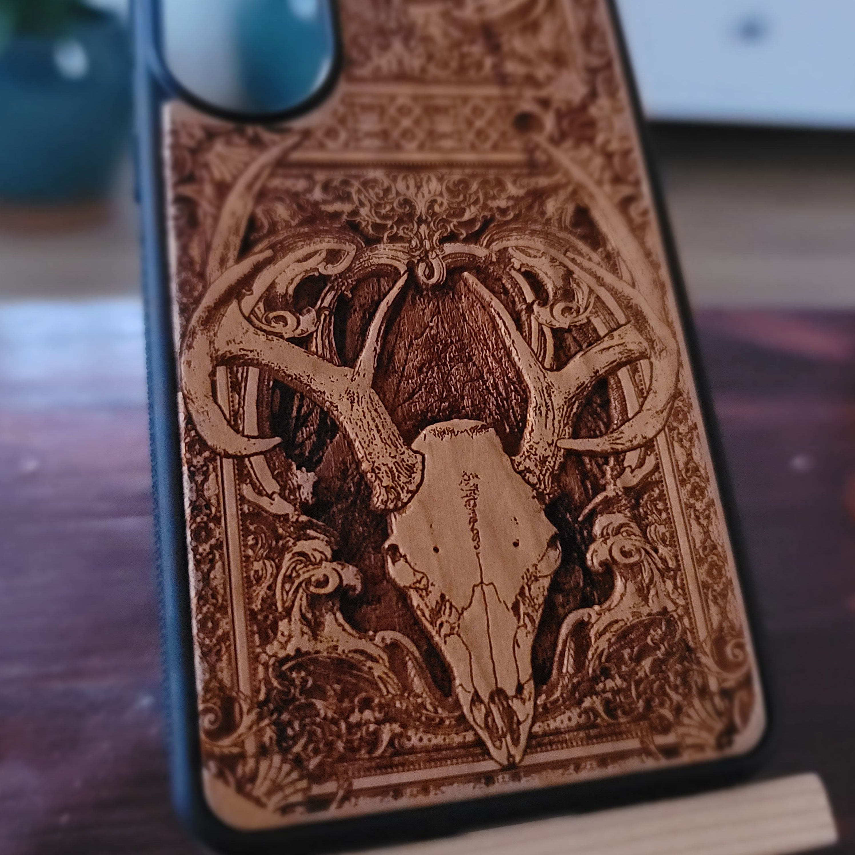 A close-up of a phone case with an intricate laser-engraved design of a deer skull and antlers on a dark wood background, resting on a wooden surface.