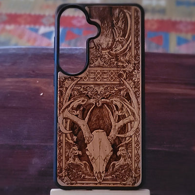 A wooden phone case with a detailed laser-burned design of a deer skull and antlers, displayed on a dark wooden surface.