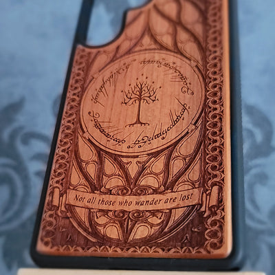 A close-up of a phone case featuring an engraved design inspired by The Lord of the Rings, with the Tree of Gondor, Elvish script, and the quote 'Not all those who wander are lost' on a dark background.