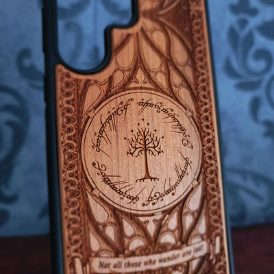 A close-up of a wooden phone case featuring an engraved circular tree design with Elvish script and the quote 'Not all those who wander are lost', shown against a blue patterned background.