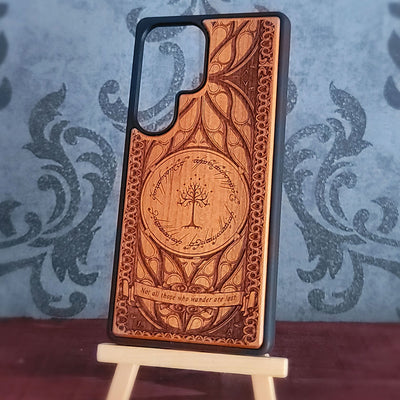 A wooden phone case with a dark gray bumper edge, featuring an engraved design inspired by The Lord of the Rings, displayed on a small wooden stand against a patterned blue-gray background.