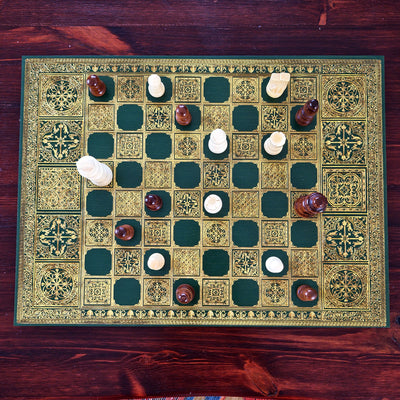 An ornate green and gold chess board set on a dark wooden table, with white and brown pieces arranged to play. The board features intricate geometric patterns and a decorative border.