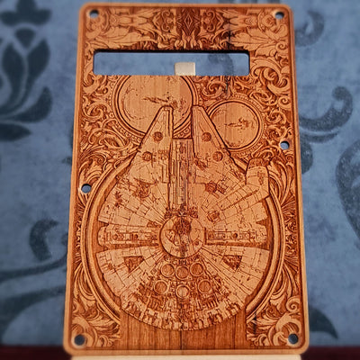 A wooden guitar backplate with a detailed laser-etched design of the mill falcon. The backplate is brown with a light wood grain and has a rectangular shape with rounded corners.