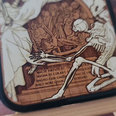 Close-up of a square wooden plaque or coaster with a dark border, featuring an engraved or burned wood illustration of a skeleton beside a figure and Latin text.