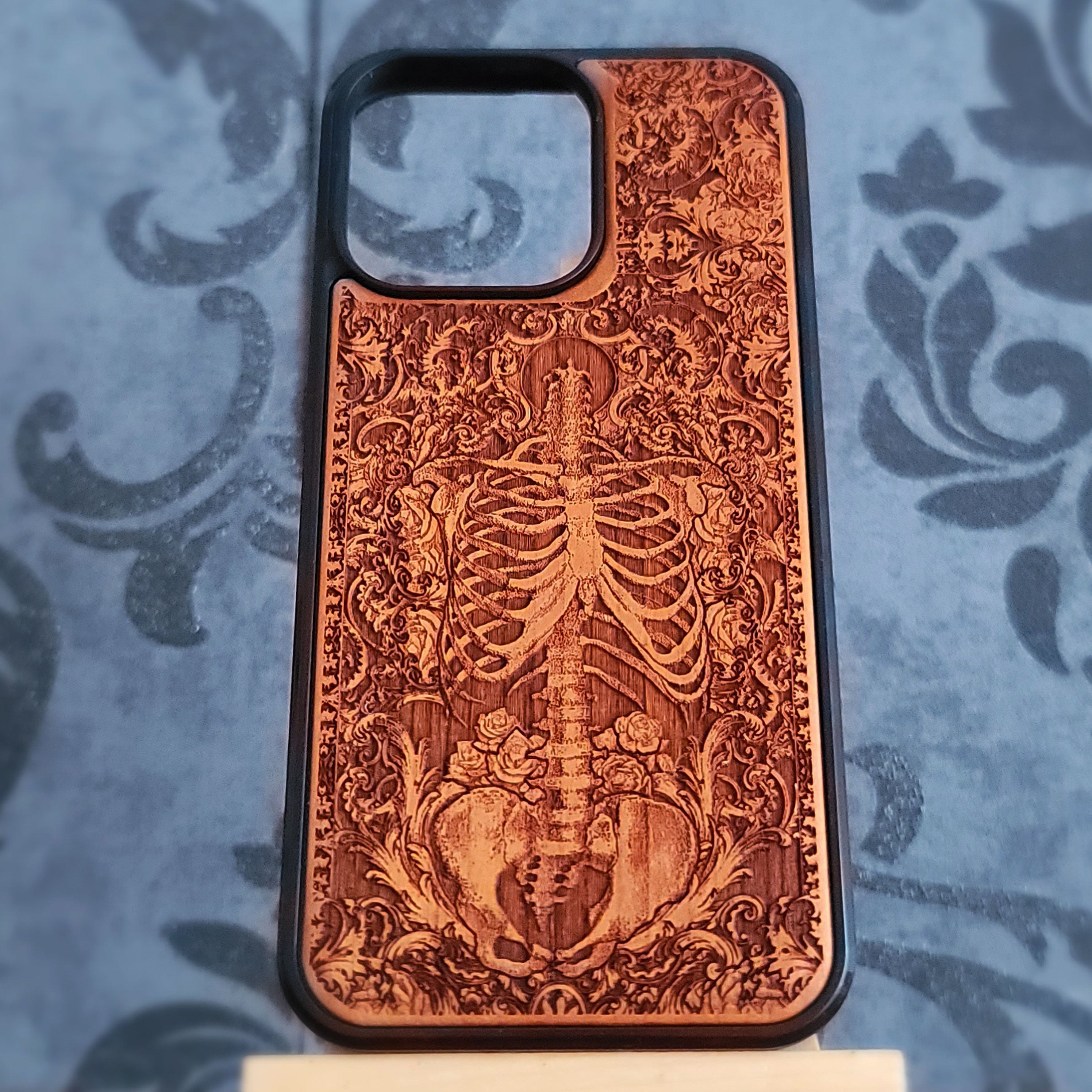 A wooden phone case with a laser-engraved skeleton and floral baroque design, featuring a black bumper edge, displayed on a blue damask-patterned surface.