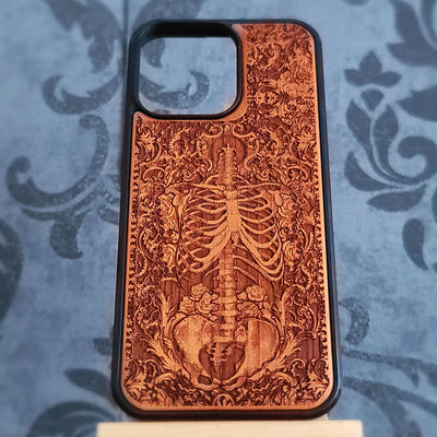 A wooden phone case with a laser-engraved skeleton and floral baroque design, featuring a black bumper edge, displayed on a blue damask-patterned surface.
