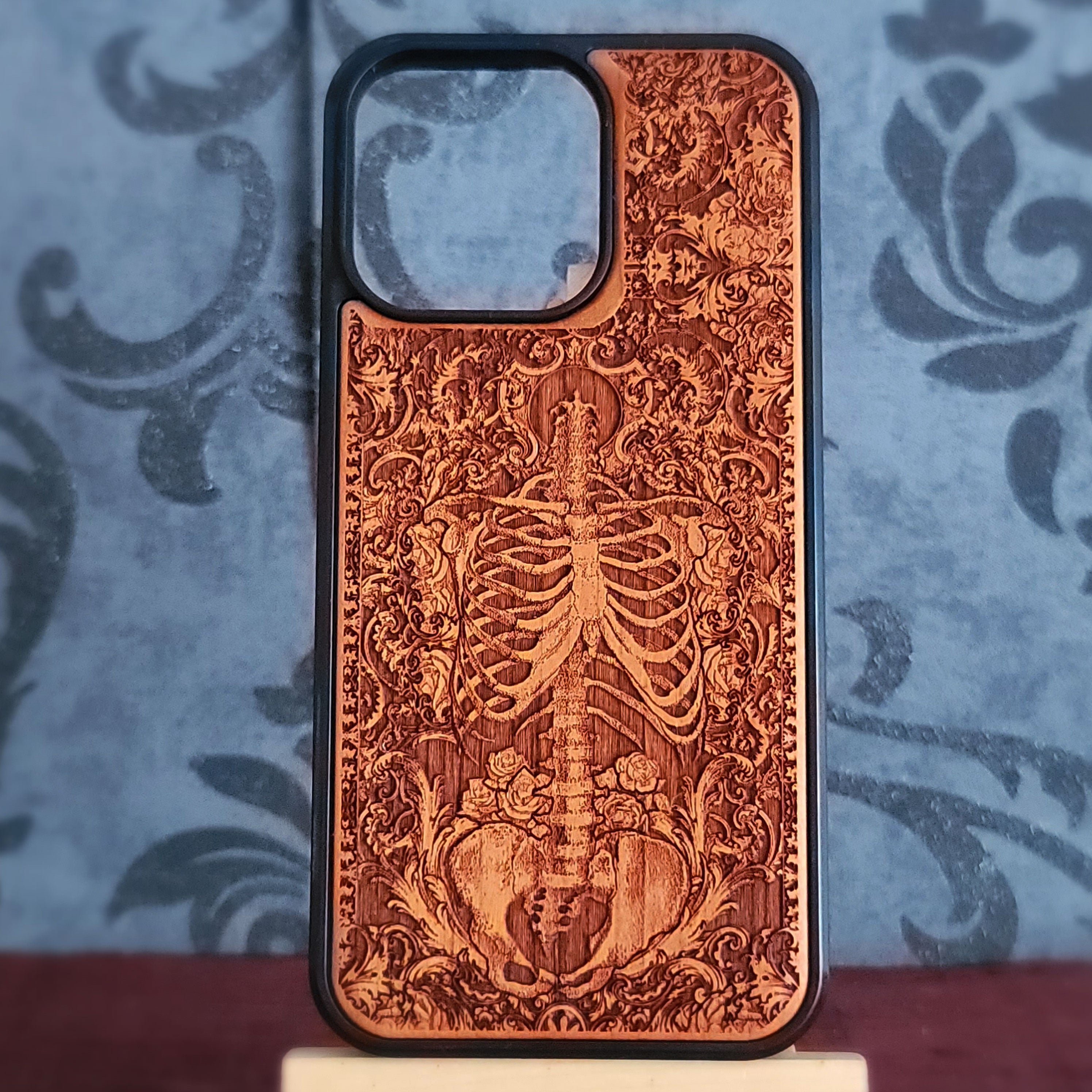 A wooden phone case with a laser-engraved skeleton and floral pattern, featuring a black protective bumper edge, displayed on a small stand against a blue damask patterned background.