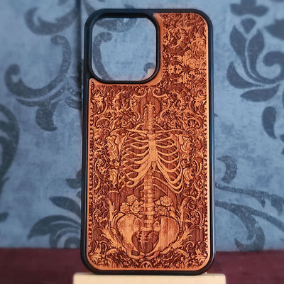 A wooden phone case with a laser-engraved skeleton and floral pattern, featuring a black protective bumper edge, displayed on a small stand against a blue damask patterned background.