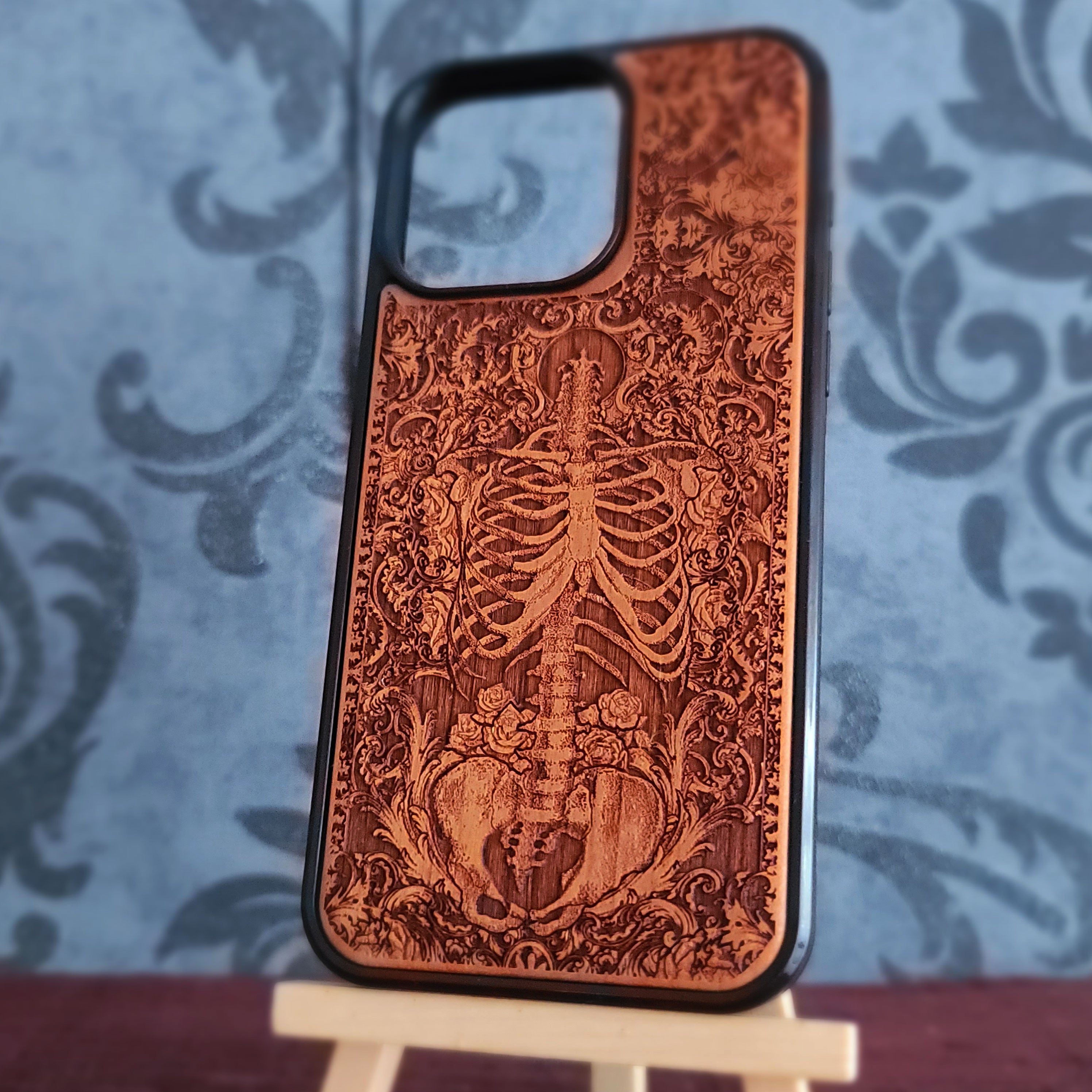 A wooden phone case with a carved skeleton and ornate scrollwork design, standing on a small display easel against a blue patterned backdrop.