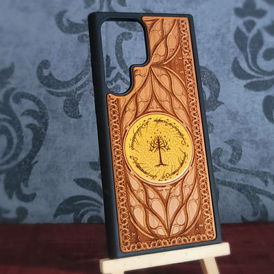 A phone case featuring an embossed design inspired by The Lord of the Rings, with a central gold Tree of Gondor emblem and a black bumper edge, displayed on a small wooden stand.