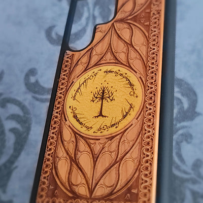 A close-up of a carved wooden phone case featuring an intricate leaf pattern and a central circular design with a tree and Elvish script, shown against a blue patterned background.
