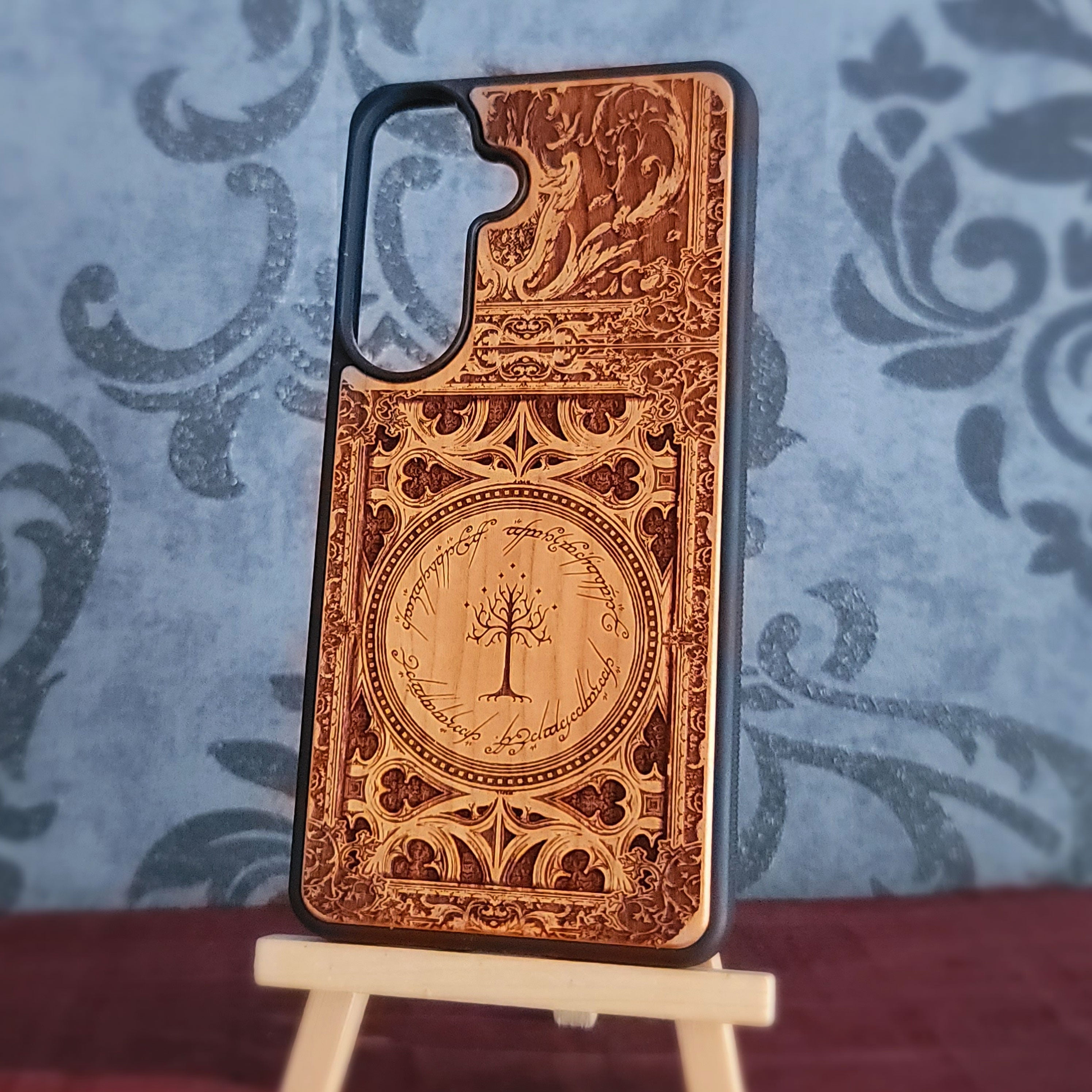 A wooden phone case with intricate laser-engraved designs and a tree motif, displayed on a small wooden stand against a blue patterned backdrop.