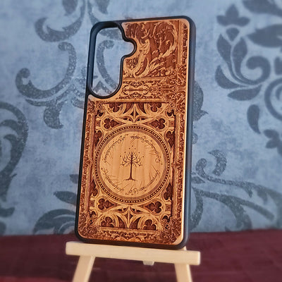 A wooden phone case with intricate laser-engraved designs and a tree motif, displayed on a small wooden stand against a blue patterned backdrop.