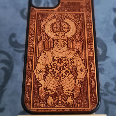 A close-up of a phone case featuring an intricate laser-engraved design of a knight in armor with a halo, set against a stained-glass-style background. The case has a warm brown wood-grain finish with a black bumper edge, 