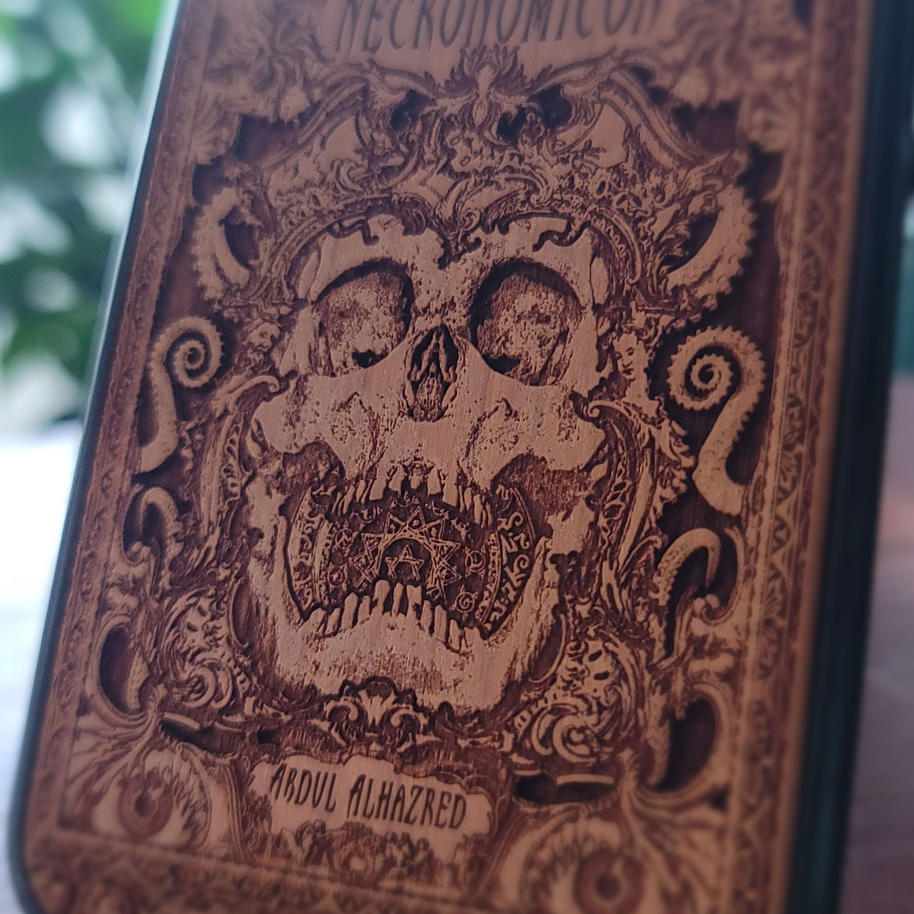 A close-up of a Necronomicon-themed book cover featuring an intricately engraved skull and ornate gothic illustration in dark brown on a tan background.