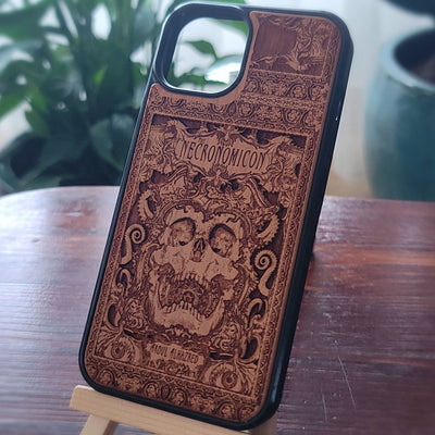 A wooden phone case with a dark gothic Necronicon skull design, displayed on a small wooden stand on a wooden table with a blurred background.