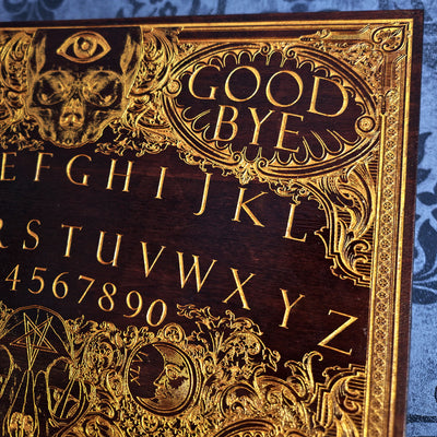 Close-up of an ornate dark wood Ouija board with raised gold lettering and decorative motifs, including the words 'GOOD BYE' and the alphabet with numbers.