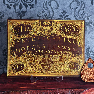 An ornate gold and dark wood Ouija board with intricate filigree details, A matching planchette rests to the right.