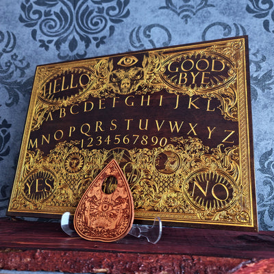 A dark wood Ouija board with ornate gold foil lettering and symbols, displayed on a clear stand with a matching teardrop-shaped copper planchette in front