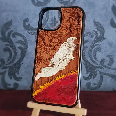 A wooden phone case with a black bumper edge, featuring a laser-engraved design of an astronaut floating above a red landscape with yellow flames, displayed on a small wooden stand.