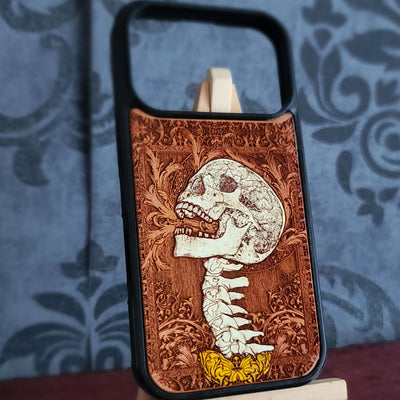 A phone case with a black bumper and a custom wooden back featuring a detailed laser-engraved skull and spine design on a brown wood grain background, displayed on a small stand.