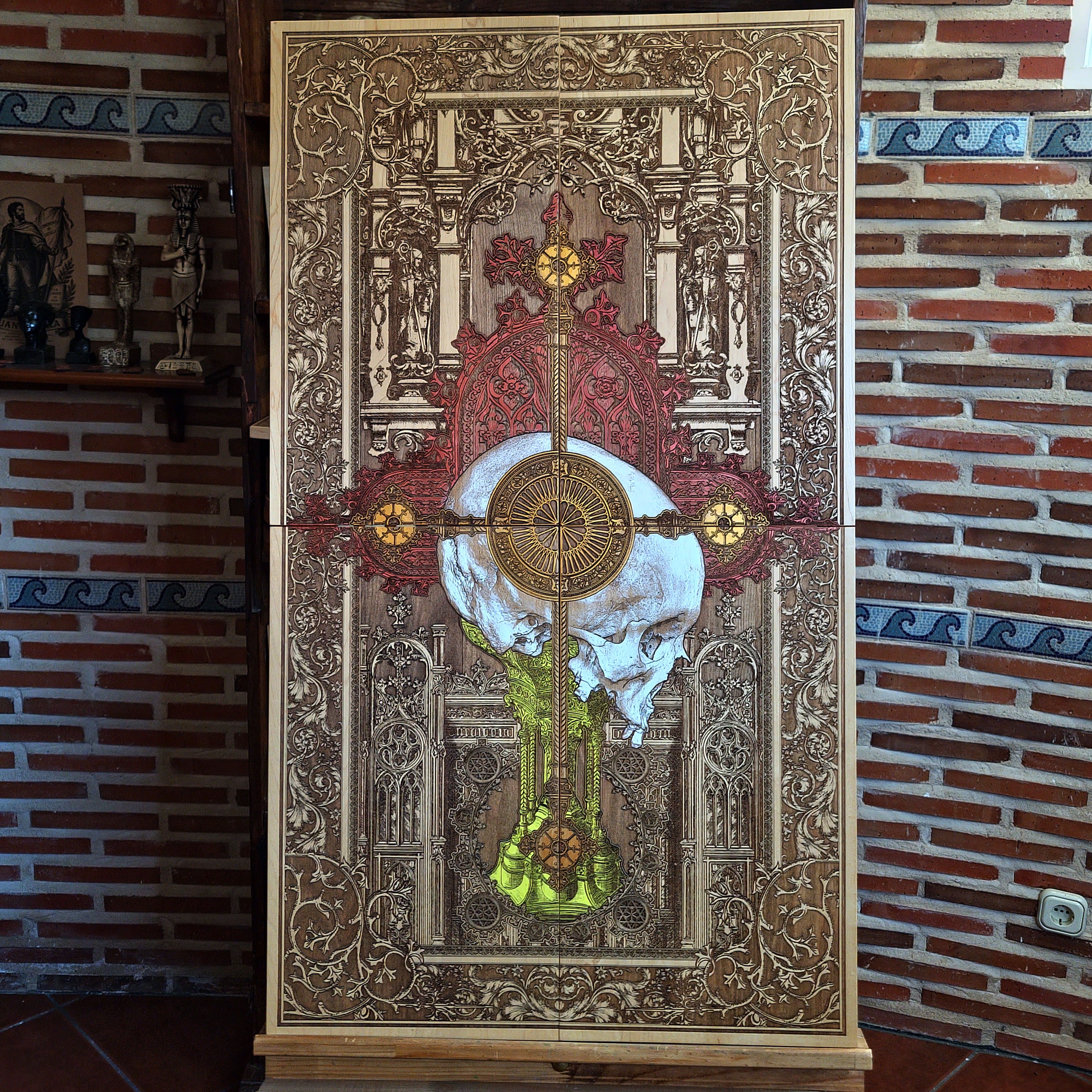 A detailed wooden artwork featuring a skull at the center, overlaid with a cross and intricate gothic architectural elements. The artwork is primarily brown with accents of gold, red, and yellow. 